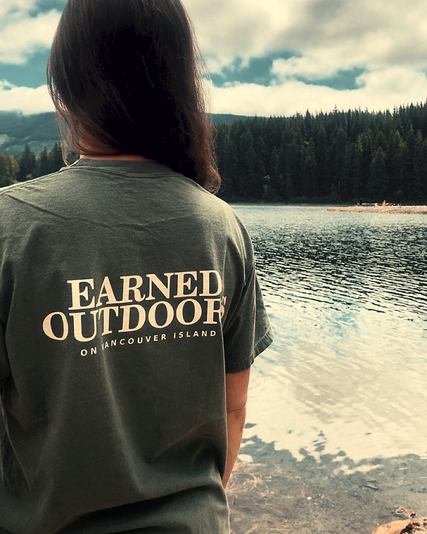 The Long Haul Tee in moss beside the lake, Earned Outdoors back print visible